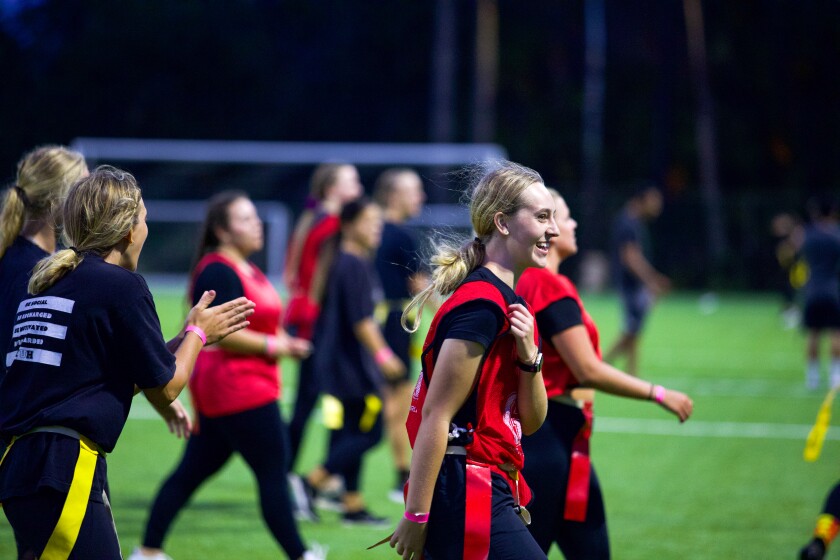 Several girls wearing black pants and black T-shirts are on the turf field walking to the right. Some are wearing yellow flag and others are wearing red flags with a red jersey. One girl is clapping.