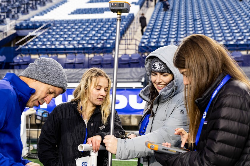 Three students stand next to a professor as the four of them take moisture measurements of the football field
