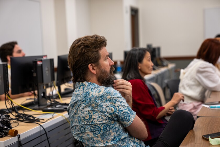 adults sit at desks and pay attention to a lecturer off screen