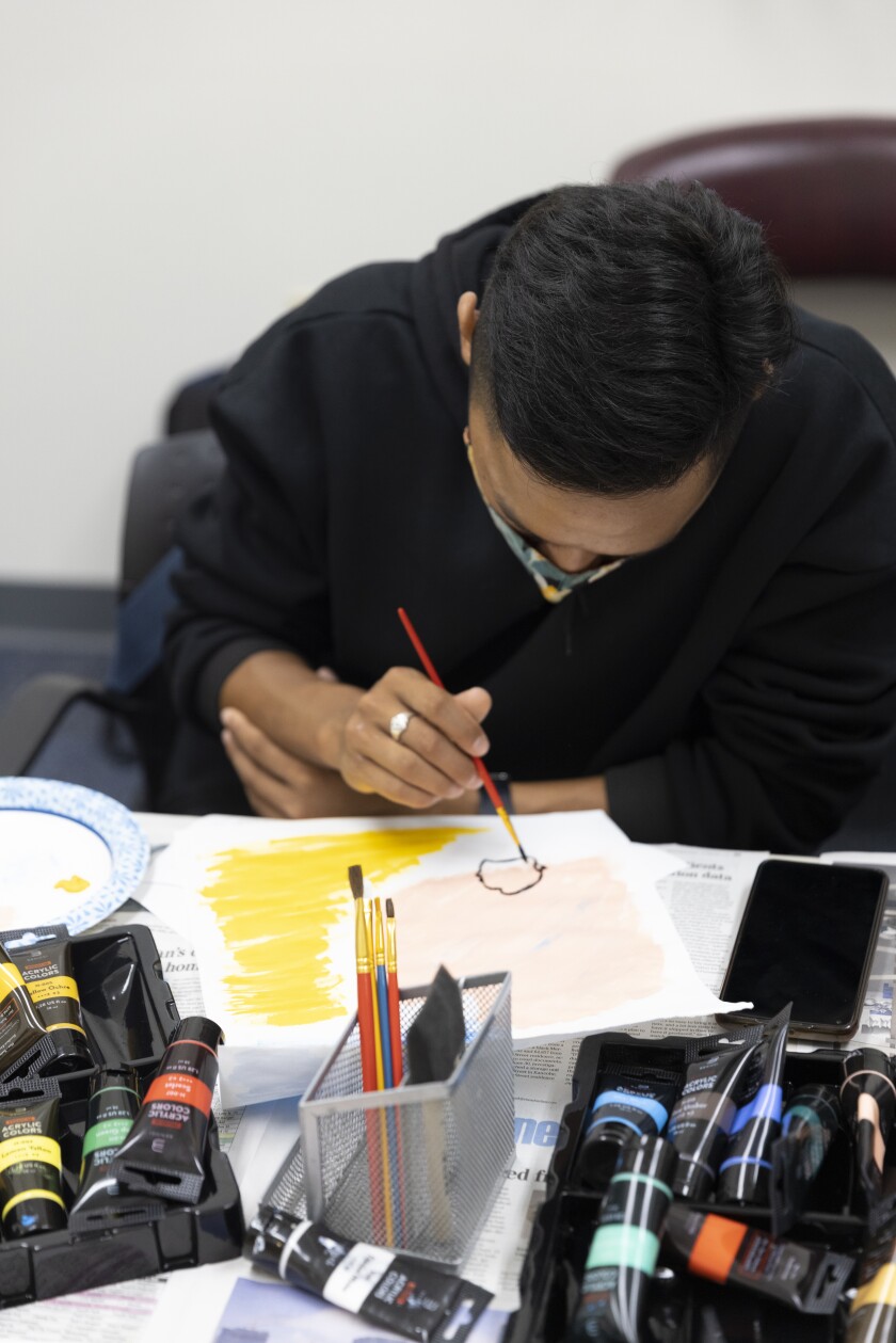 Student in black hoodie and wearing a silver ring is seated, surrounded by paint tubes, newspaper, paintbrushes and a cell phone on the desk in front of him. He is holding a red paintbrush and painting a shape on a pale pink and yellow paper.