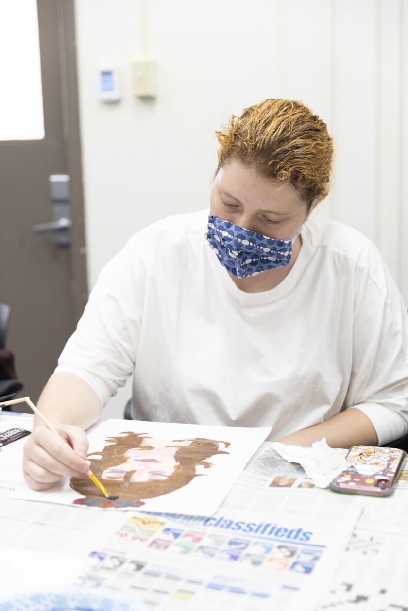 Female student wearing plain white shirt and wearing a blue patterned mask is seated, painting a portrait of a woman with brown hair