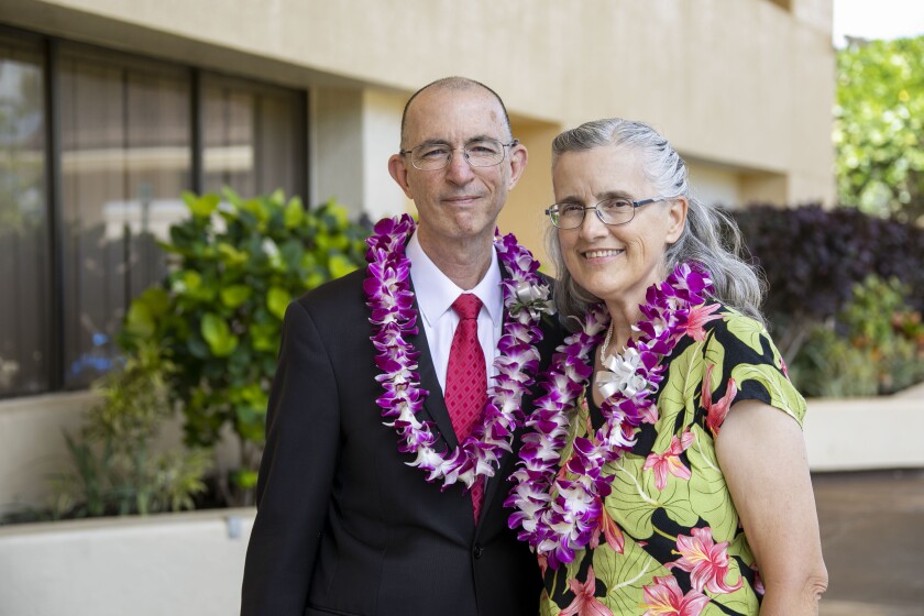 John and Rhonda Bell together wearing purple and white flower leis wearing a suit and black, green and pink blouse with an off-white building behind them.
