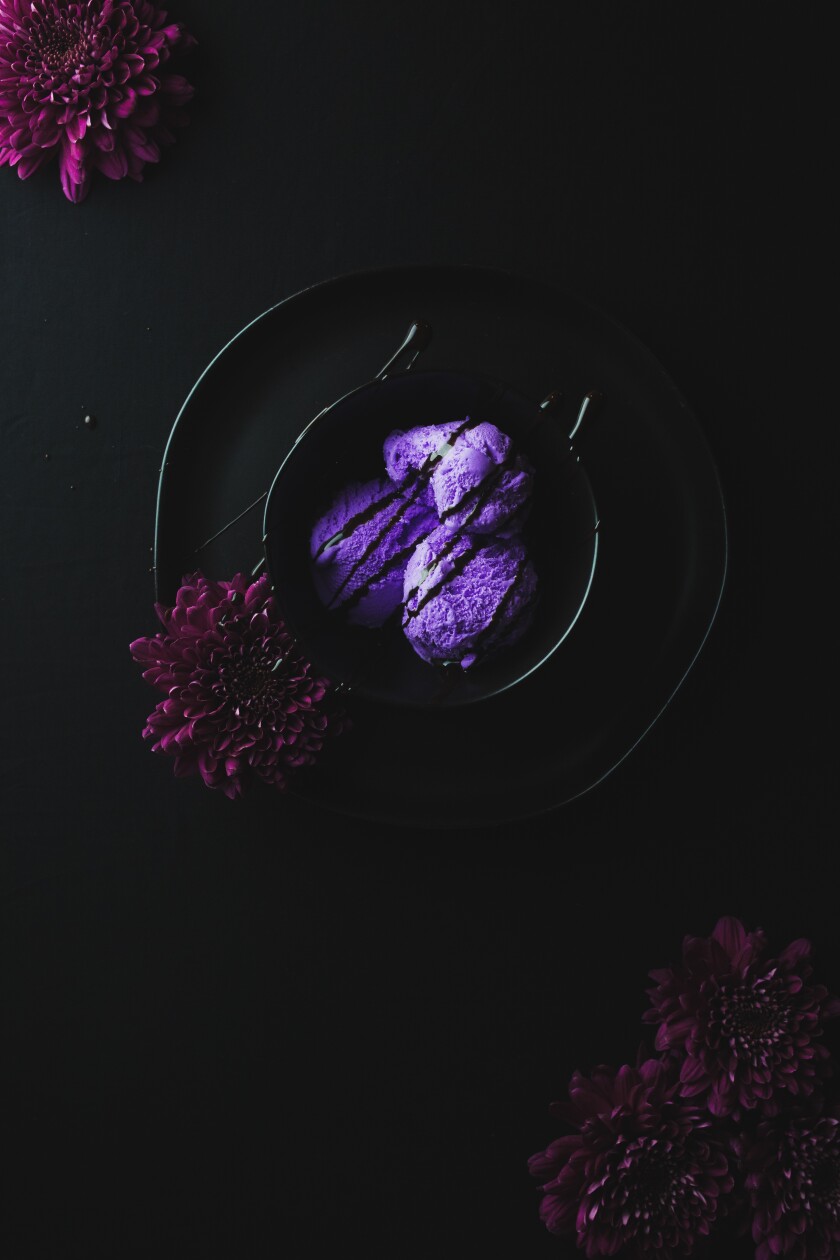 Ube ice cream in a black bowl on a black plate with chocolate syrup drizzled over the top. There are dark purple flowers in the right corner, left corner and on the left side of the bowl. The photo has a black background.
