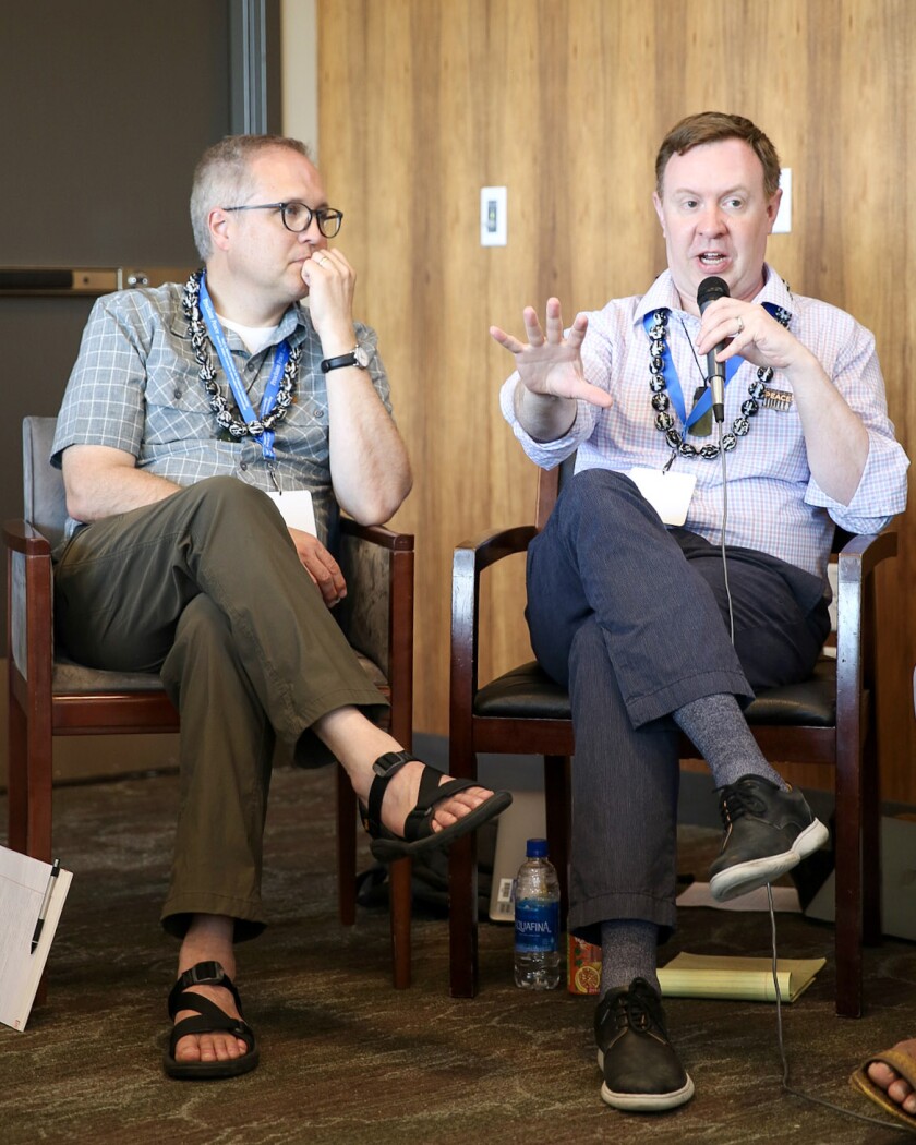 Two men are seated one wearing a plaid shirt and blue pants and talking into a microphone, and the other is wearing a gray plaid shirt, brown pants and sandals.