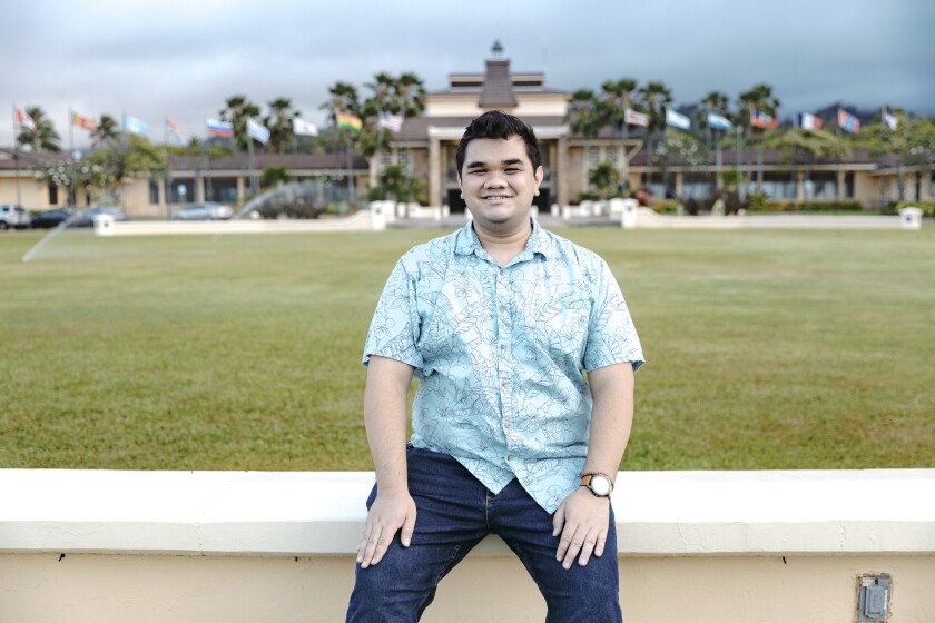 Burgess smiling while wearing a blue aloha shirt and black jeans sitting down with a field and the front of the BYUH campus buildings and flags behind him.
