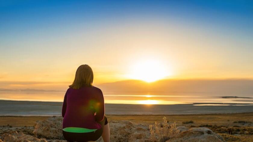 A woman sits on the shore and stares off into the distance at a sunset