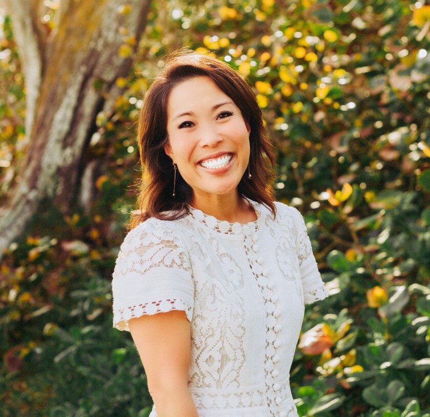 Jennifer Kajiyama Tinkham wears a white dress and stands in front of greenery outdoors.