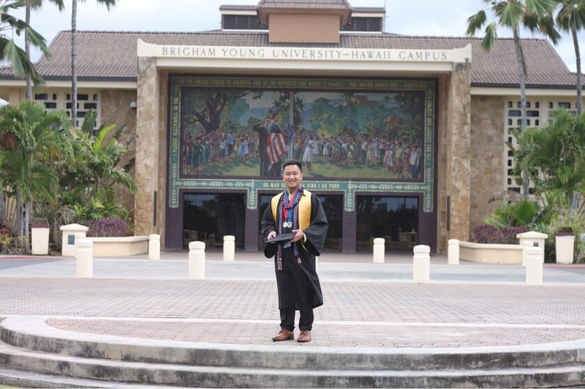 A young man in a black graduation gown and holding his graduation cap, stands in front of the McKay mural at BYU-Hawaii.