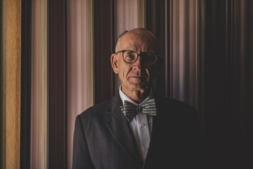 Anderson looking at the camera, wearing a black suit with a black-spotted bowtie and glasses with a purple wall behind him.