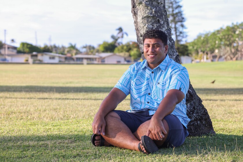 Kelvin Uili sitting down on the grass and posing for a photo with a coconut tree behind him
