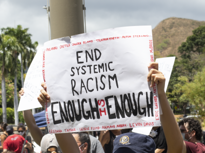 Someone holding up sign saying "End systemic racism enough is enough" with the names of black lives matter victims on the side. In the background, there is a crowd of people and palm trees and mountains.