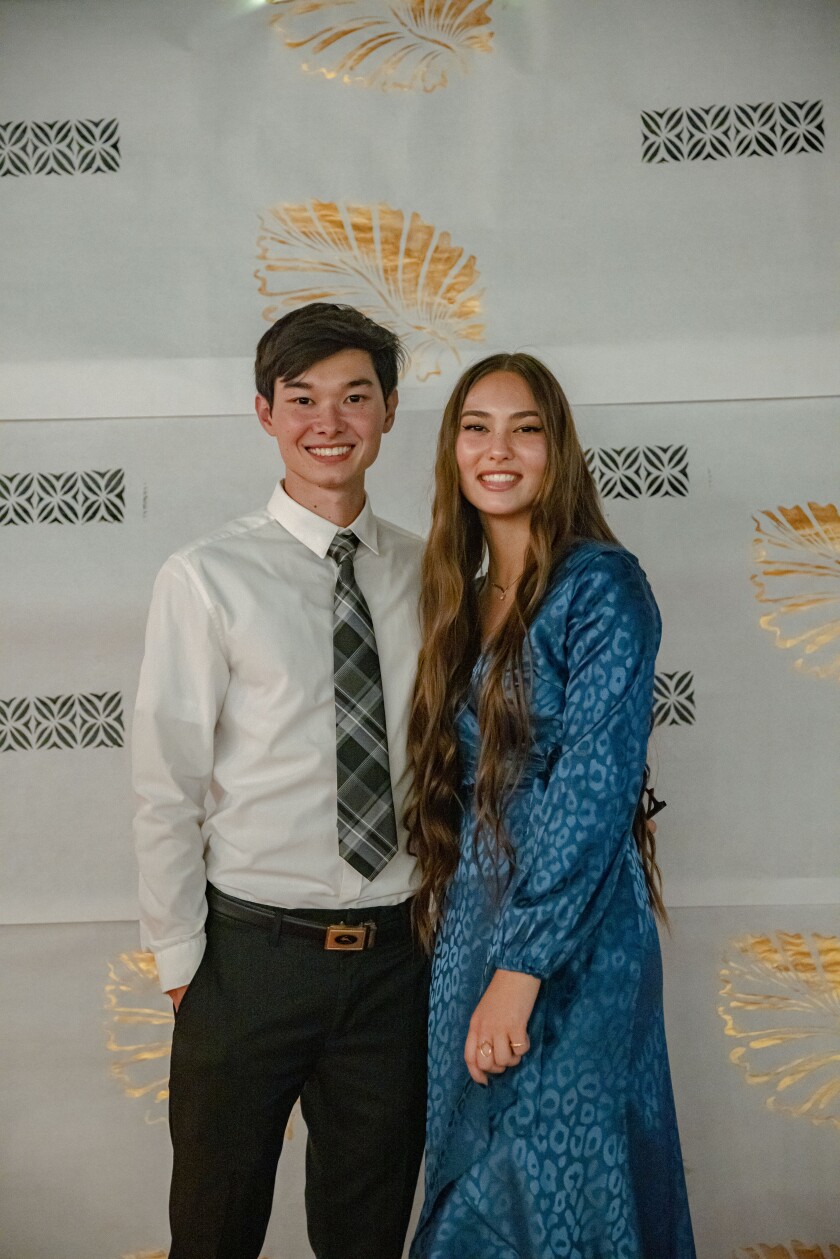 A couple pose together in front of the same white, gold and black backdrop. The boy is wearing a white long-sleeve button-up, black pants, grey, black and white plaid tie, and a brown belt. The girl is wearing a long-sleeve blue cheetah-print dress.