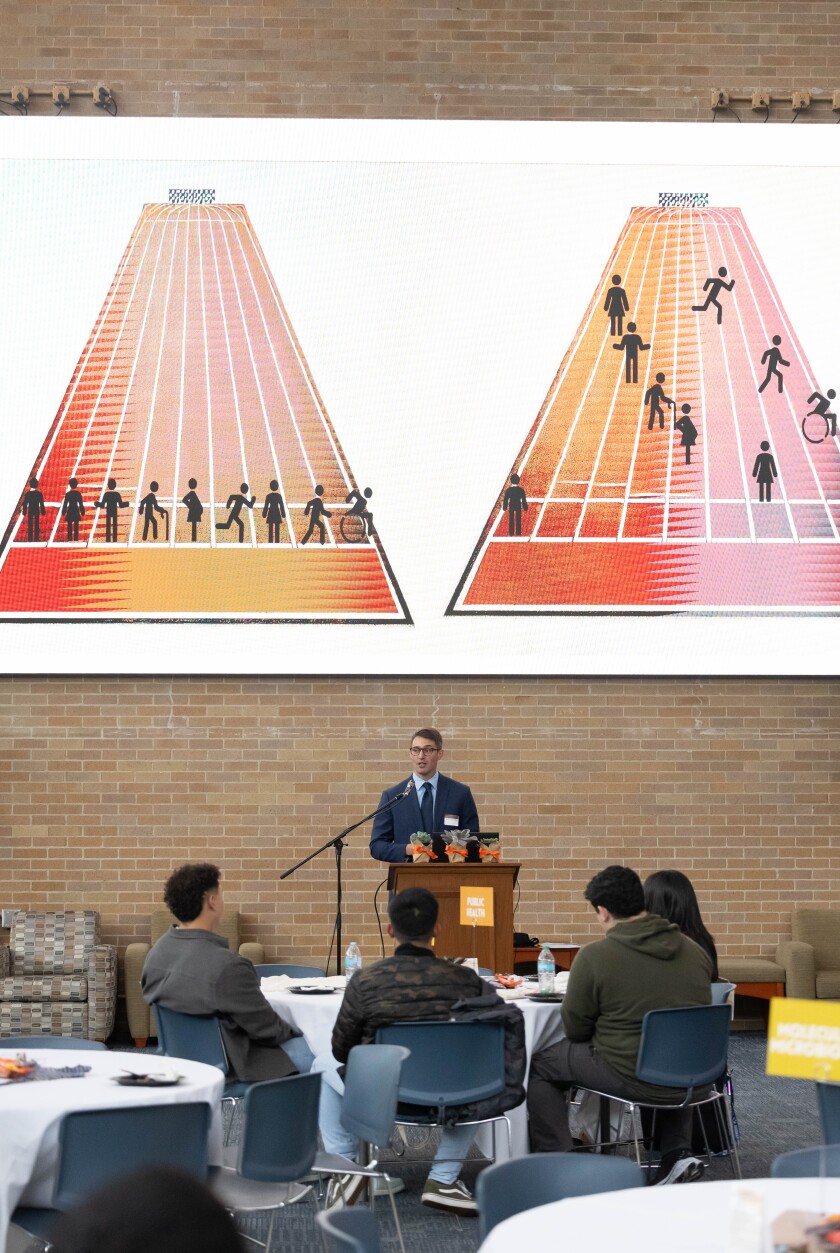 A man wearing a suit and glasses standing behind a podium and addressing the students infant of him. Projected on the screen behind him is a visual depiction of the $100 race and the social inequalities among students.