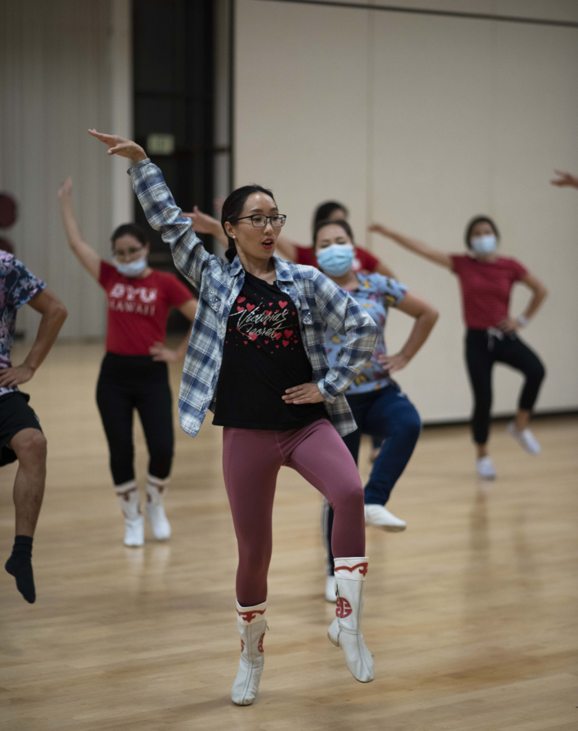 Narantsatsral Ganzorigt dancing with her left leg up and her left hand on her hip and other hand up in the air wearing white and red boots, purple leggings, black shirt and white and blue plaid shirt with other dancers behind her.
