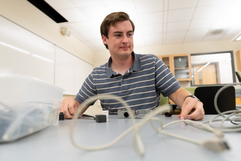 A male student in a striped gray polo messes with a computer charging cord plugging it into a measuring machine.
