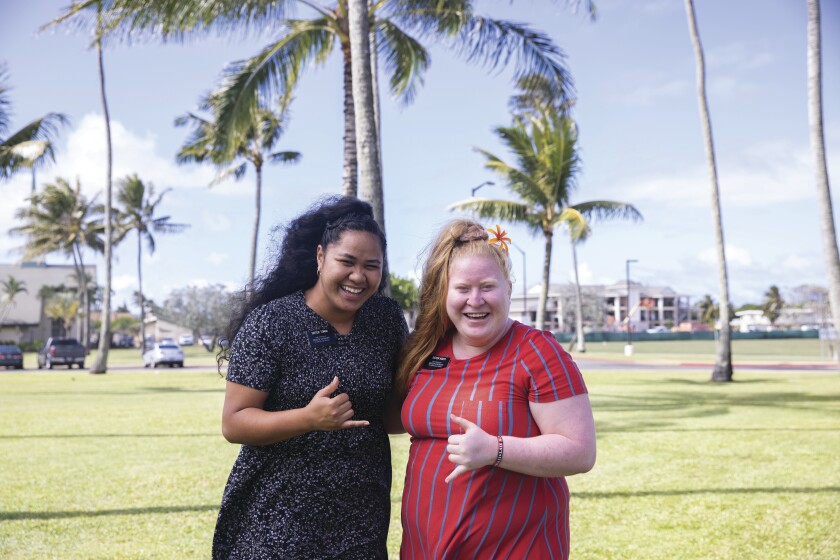 landscape shot of two sister missionaries holding up shakas and smiling on BYUH grounds with green grass and palm trees behind them