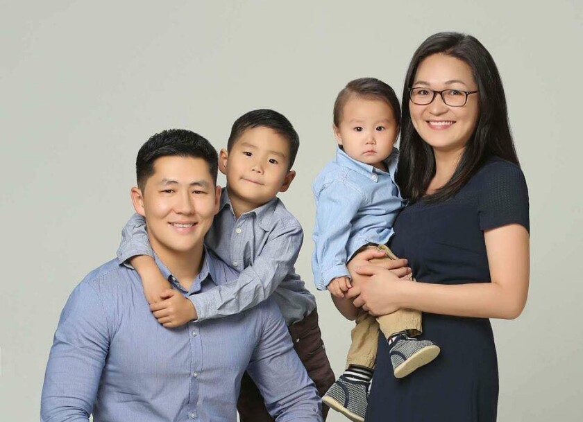 Gankhuyag Tsogoo (left) sits with his sons arms around his neck and his wife on his right side holding his younger son, the boys wearing blue button up shirts and his wife wearing a dark blue dress with a grey/white wall behind them.
