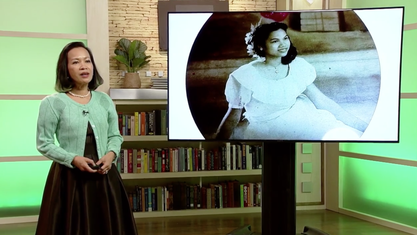 Screenshot of Dr. Astrid Tuminez wearing a brown dress and green cardigan with a screen of a photo of her family wearing all white on it.