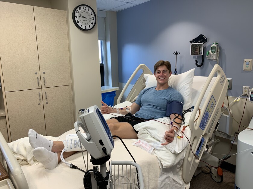 A man lies propped up in a hospital bed smiling and holding both hands in shaka signs, the pinkie and thumb extended out and the other fingers curled over. His arms are hooked up to a machine and blood through the plastic tubes. He has light-toned skin, middle-parted short brown hair, a light blue tshirt and black shorts.