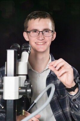 A pale man with brown hair and glasses wearing a plaid shirt smiles in front of a microscope