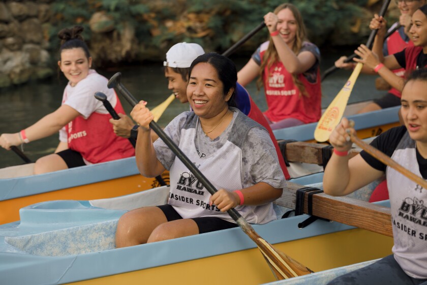 Six female students rowing a canoe at the Polynesian Cultural Center. They are all wearing dark colored shirts, T-shirts and red and white BYUH Seasider Sports jerseys.