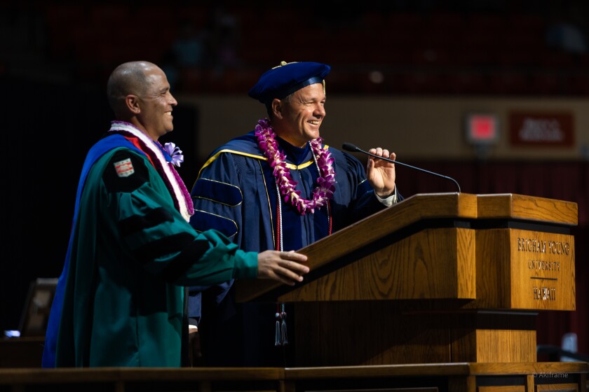 President John S. K. Kauwe III and Vice President of Academics, Isaiah Walker, stand together at the pulpit. President Kauwe is wearing a green robe, with a blue sash and a purple and white lei. Vice President Walker is wearing a purple and white lei with red and white ropes and a blue and gold robe. He also has a blue and gold hat on. They are both smiling.