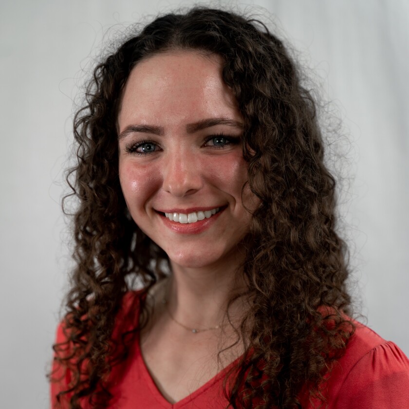 Katelyn Bell is a young woman with dark curly, shoulder-length hair. She is wearing a red blouse and a small gold necklace.