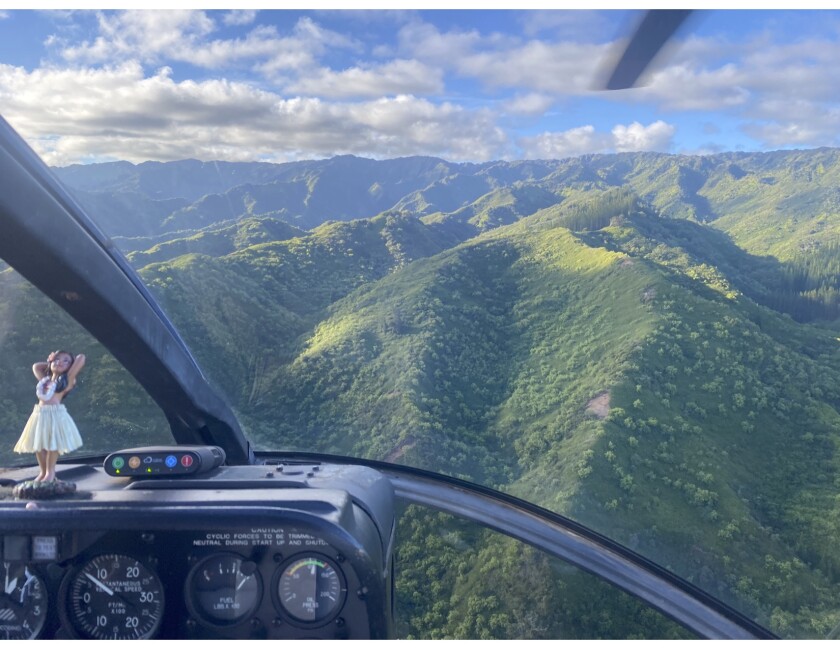 A helicopter view of green Hawaiian mountains. In the left corner, a small hula figurine can be seen sitting on the dashboard of the helicopter.