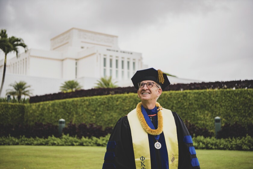 Anderson looking up and smiling wearing a graduation cap and gown with a yellow sash, yellow lei and blue medal around his neck with the Laie Hawaii Temple behind him and greenery.