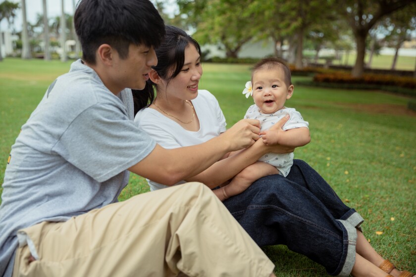A couple holds there baby while sitting on grass with palm trees in the background.