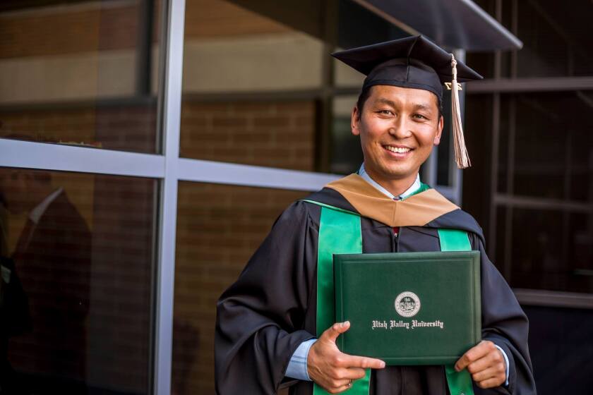Tserennyam Sukhabaatar wearing a black cap and gown with green and brown on it holding a green folder that says "Utah Valley University" with a black-mirrored wall behind him.