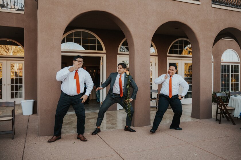Three men wearing dark grey/black pants, brown shoes, white button-up shirts and dark orange/red ties dancing in front of a building with arches. The man in the middle is also wearing a suit coat and a ti leaf lei around his neck.