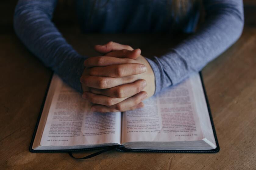 A woman's arms and hands are visible. She is wearing a light blue long-sleeve shirt. Her hands are clasped in prayer over open scriptures set on a wooden table.