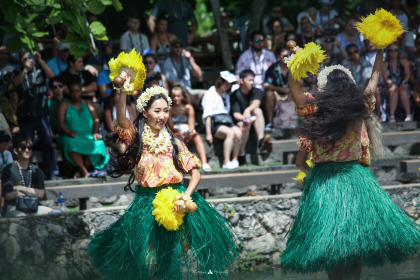 Ganzorigt dancing in a grass skirt, orange and yellow shirt and flower leis around her neck and head holding up wooden handles with yellow feathers on them with a crowd behind her.