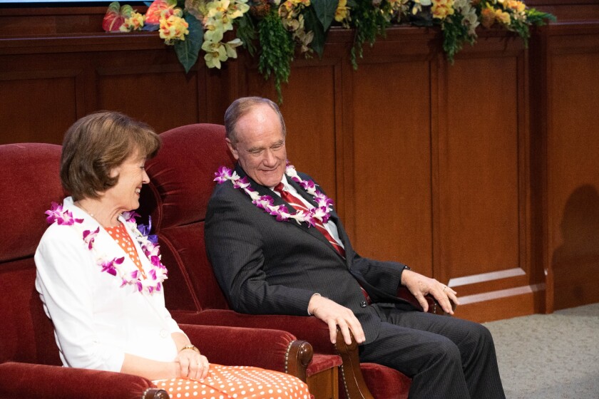 Wearing flower leis, John and Susan Tanner sit beside each other on red chairs.
