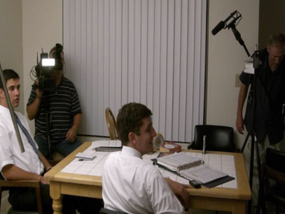 Two missionaries sit around a table with a camera crew