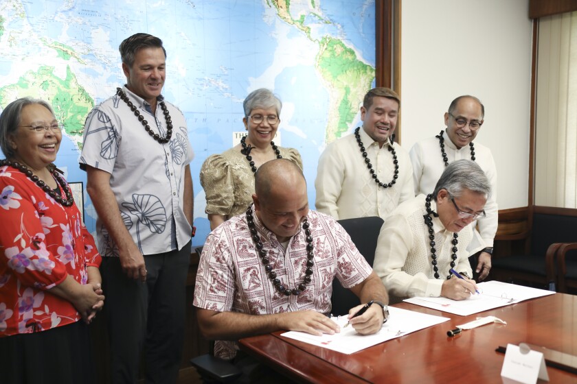 Two persons signing contracts with other persons standing behind them watching. Everyone is smiling.