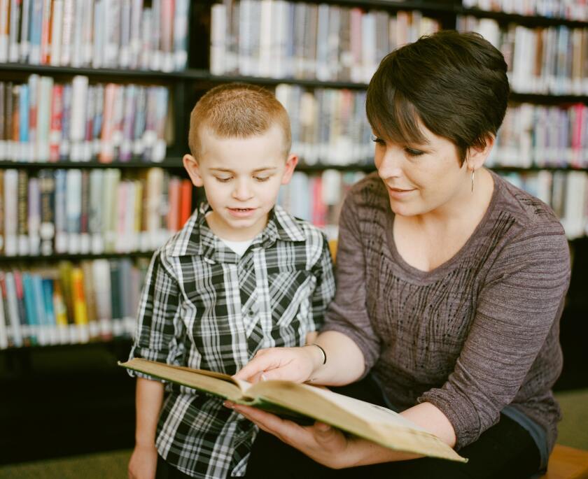 A woman dressed in a brown top reads a book in a library with a boy wearing a plaid shirt