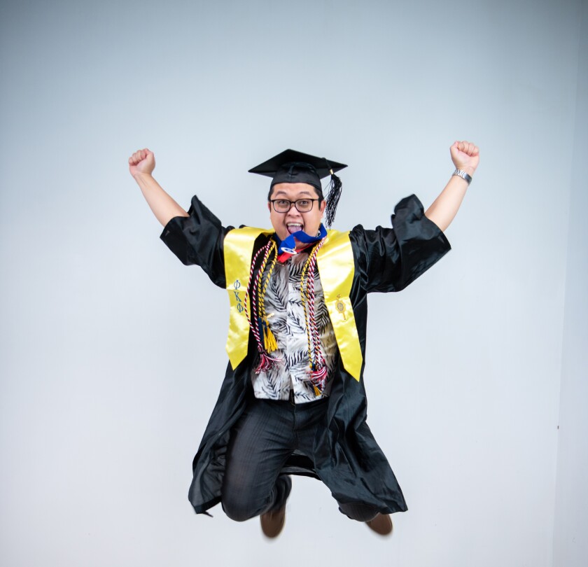 Dela Peña smiling jumping in the air wearing his graduation cap and gown and a yellow sash