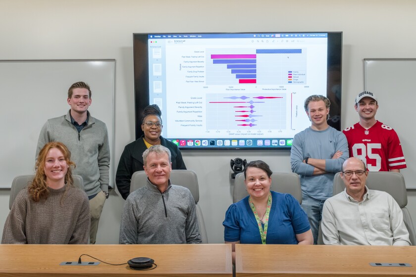 A group of researchers sits together and smiles. Behind them is a screen with science on it.