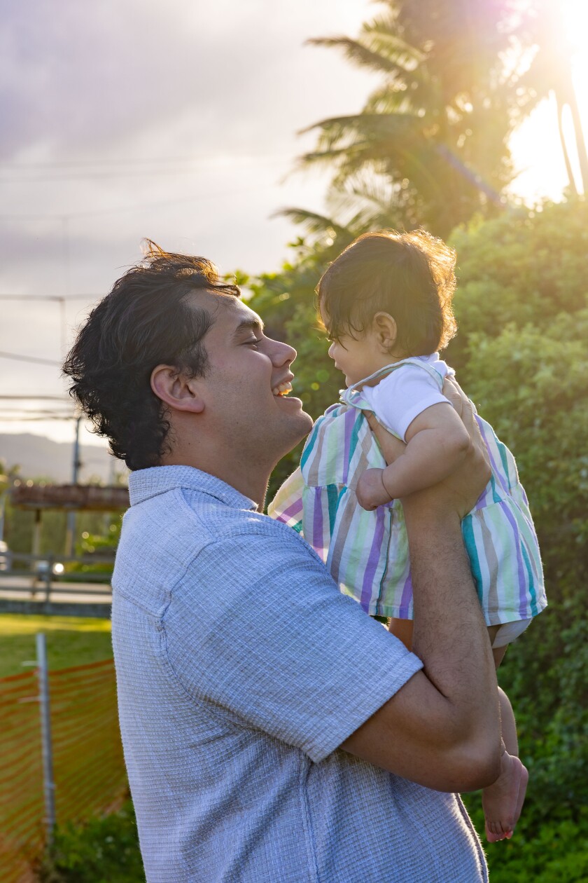 Daniel Hootini smiles at his daughter as he lifts her into the air during a golden hour moment outdoors.