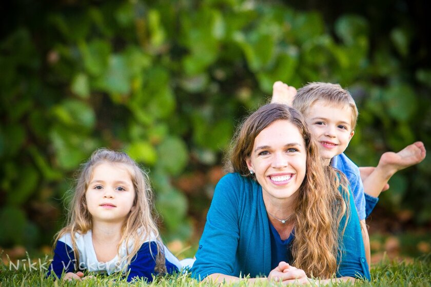 Shenley Puterbaugh lays on the grass smiling with her son on her back and her daughter on the left of her, all wearing blue