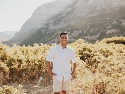 A man wearing a white, short-sleeved, button-down shirt stands in front a fall foliage with mountains in the background.