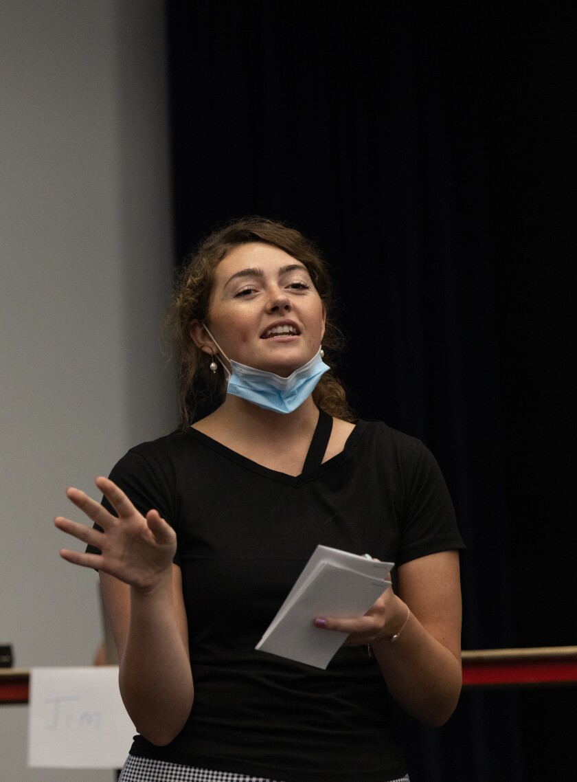 Emarie Majors, wearing a black shirt and black and white checkered pants, stands in front of the crowd in the Little Theater to address them.