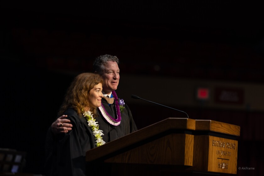 Elder Jeremy R. Jaggi and Sister Amy S. Jaggi standing at the pulpit in the Cannon Activities Center. They are both wearing black graduation gowns. Amy Jaggi is wearing a green and white lei and Jeremy Jaggi is wearing a purple and white lei and blue tie.