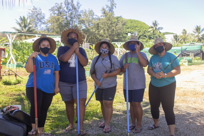 Five Samoan students stand together in t-shirts, leggings, shorts and hats holding rakes or shovels with a garden behind them.