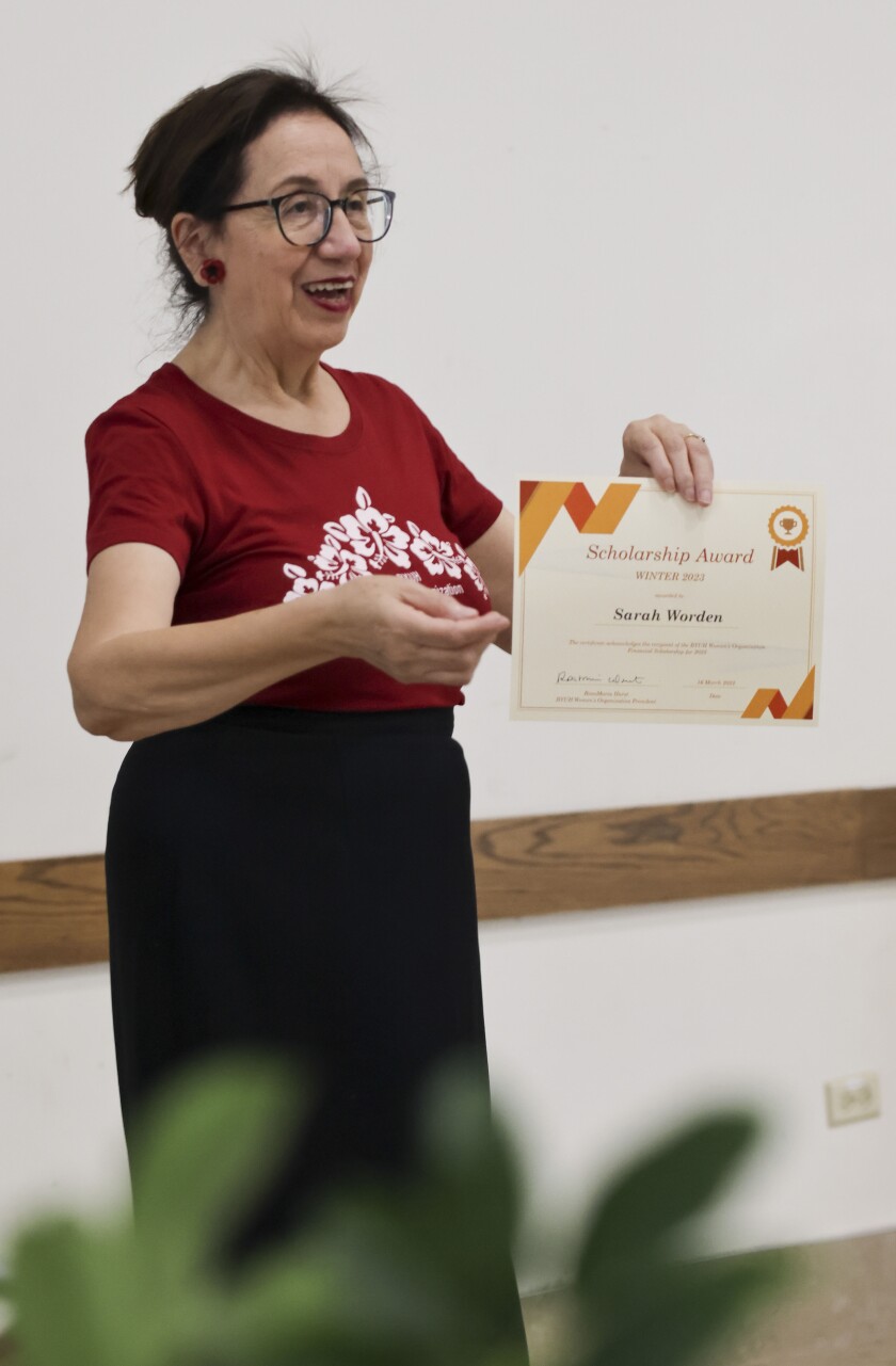 A woman dressed in a red top and black skirt holds up a certificate for one of the BYUH Women's Organization scholarship recipients.