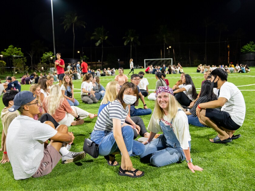 landscape shot of a crowd of students sitting on the soccer field in circles, with two female students facing the camera and smiling