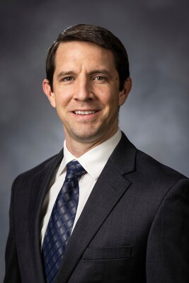 Seth Taylor. A light skinned, dark brown haired man wearing a black suit and metallic blue tie, with a gray background, is smiling in a portrait style photo.