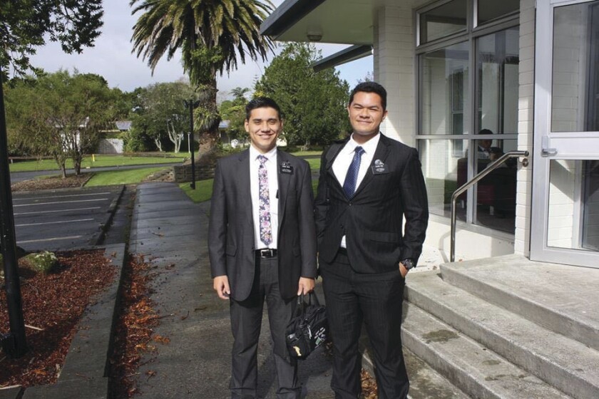 Two missionaries wearing suits, ties and missionary names tags, stand outside of a church building in New Zealand.